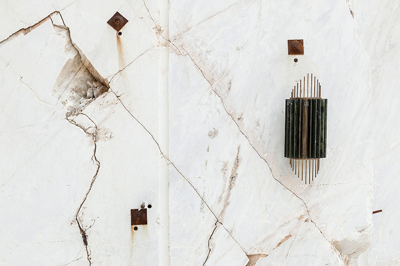 OnEntropy sculptural wall-mounted light fixture in dark green marble and brass, installed on raw white marble quarry surface with visible fractures and rusted mounting elements