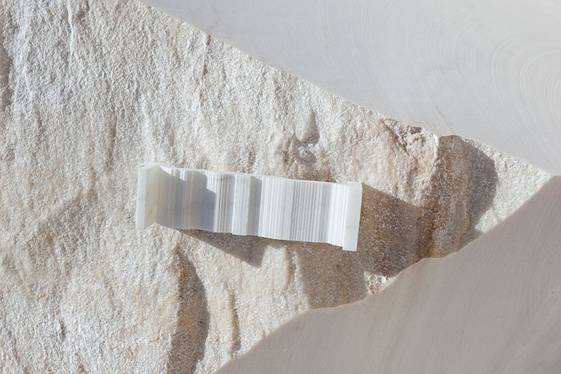 Sculptural white marble object by OnEntropy with fine vertical ridges, placed on a textured quarry stone surface in soft natural light.