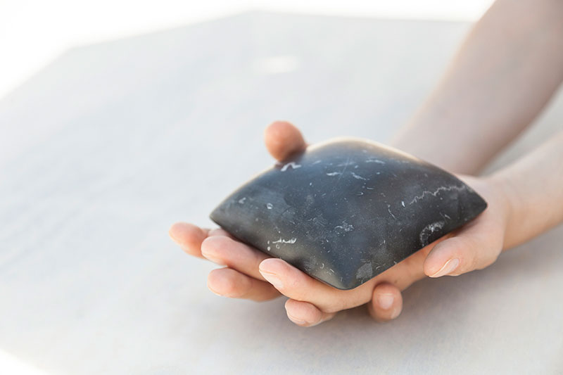 Hands gently holding a smooth, polished black marble object by OnEntropy, shaped like a soft cushion, showcasing tactile quality and sculptural craftsmanship.
