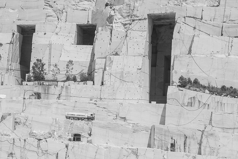 Black and white photo of a large marble quarry with vertical geometric cuts, a transport truck, and visible extraction lines—illustrating the raw origin of stone used in OnEntropy's work.