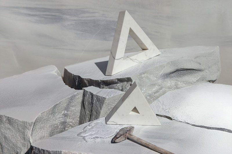 Geometric marble sculptures in triangular form by OnEntropy, displayed on raw quarried stone with chisel marks and a wooden-handled hammer, reflecting artisanal craftsmanship.