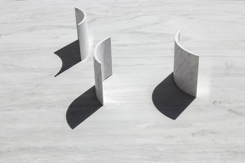 Minimal curved marble sculptures by OnEntropy casting strong shadows on a light stone surface, showcasing geometric purity and spatial rhythm.