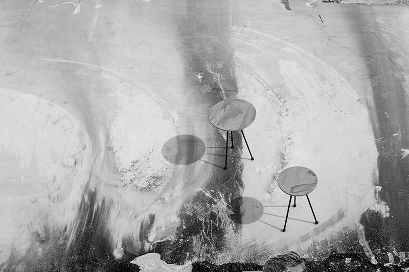 Black and white photograph of two circular marble-top tables by OnEntropy with metal legs, casting soft shadows on a textured quarry floor with sweeping surface marks