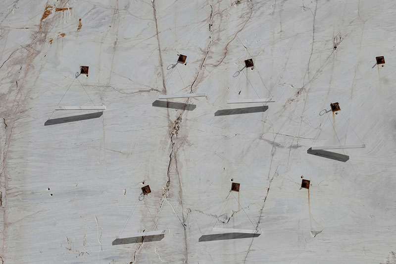 Wall-mounted minimalist marble light fixtures by OnEntropy, suspended on a raw quarry wall with visible cracks and rusted brackets, casting diagonal shadows in natural light.