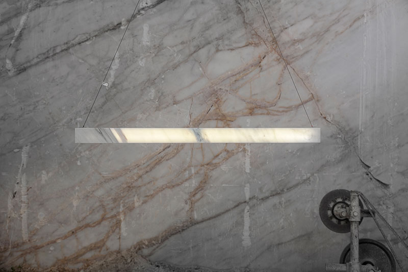 Minimal linear pendant light by OnEntropy, crafted from translucent marble and suspended in a quarry setting with visible cutting machinery and veined stone backdrop