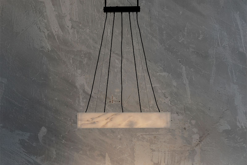 Suspended linear marble pendant light by OnEntropy, supported by multiple black cords and glowing against a textured stone wall in a refined industrial setting.