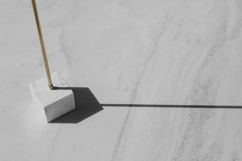 Minimalist design object by OnEntropy featuring a brass rod anchored in a white marble cube, casting a long sharp-edged shadow on a smooth light stone surface