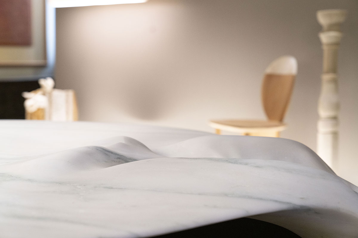 Close-up of a sculpted white marble tabletop by OnEntropy, featuring fluid, organic contours with blurred background elements including wooden furniture and soft lighting.