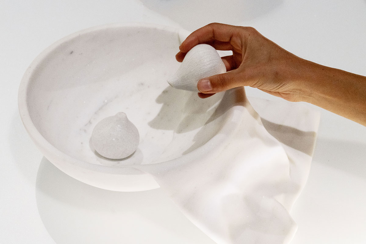 Hand interacting with sculpted white marble bowl and fig-like forms by OnEntropy, showcasing delicate craftsmanship and fluid stone drapery.