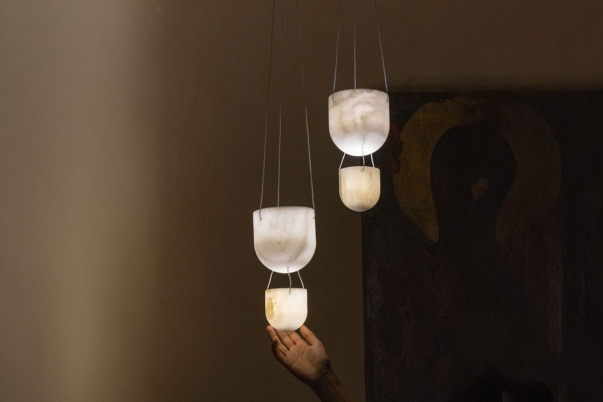 Hand reaching towards a trio of illuminated alabaster pendant lights suspended in a dimly lit interior, designed by OnEntropy.
