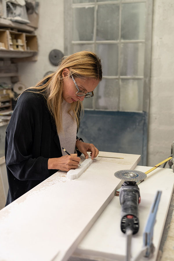 Designer working by hand on a marble prototype in the OnEntropy studio, using pencil and sculpting tools to shape fine details on a white stone surface