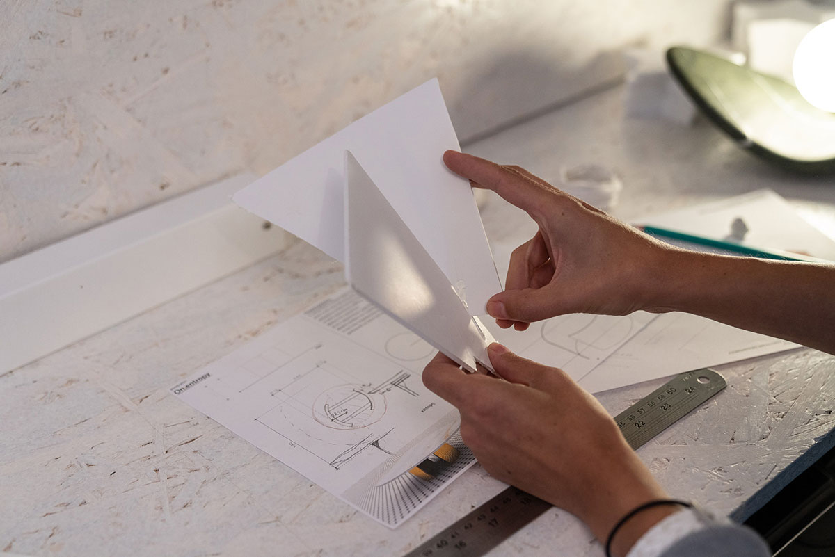 Hands assembling a white paper model of a design prototype over technical drawings, illustrating OnEntropy’s conceptual development and transition from sketch to sculptural form.