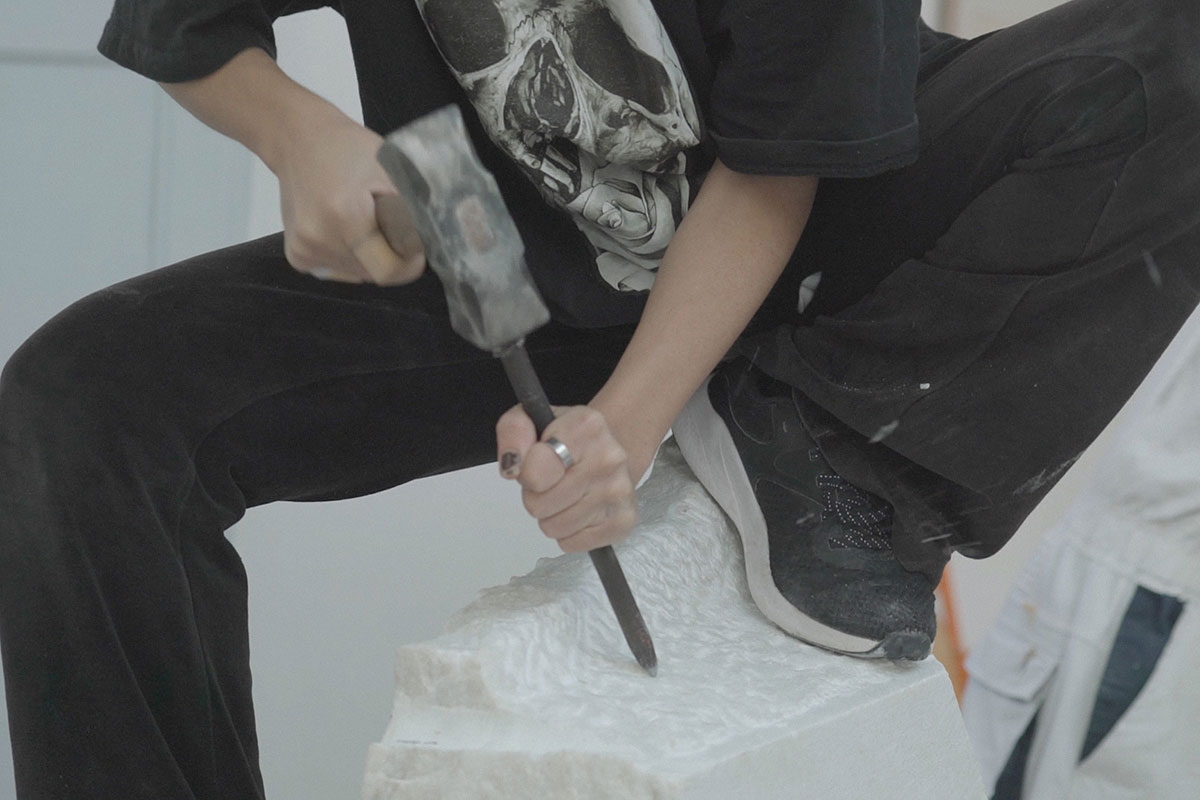 Artisan chiseling white marble while kneeling on the stone block, wearing black clothing and working with focused physicality—capturing OnEntropy’s raw and expressive craftsmanship.