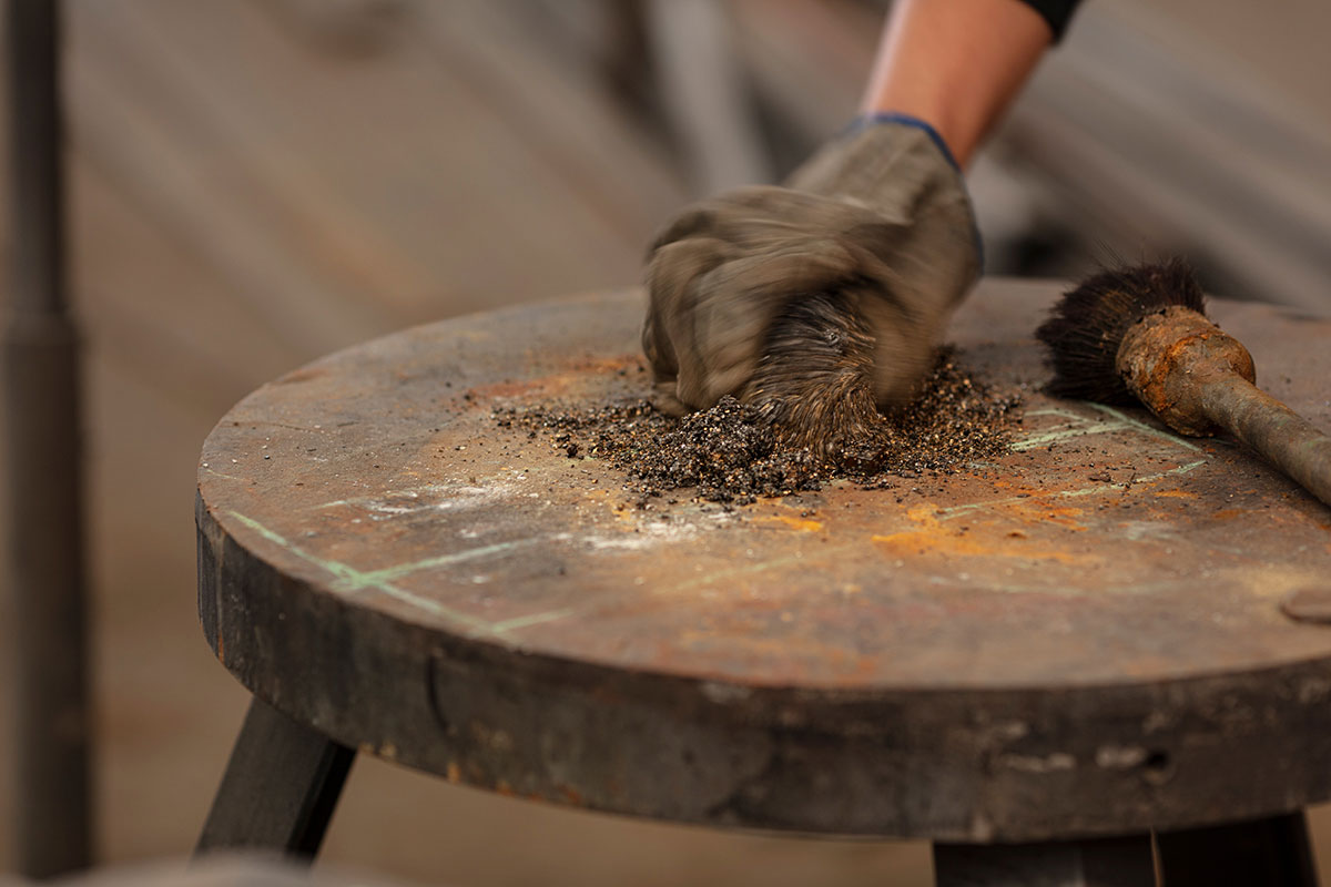 Gloved hand scrubbing metal filings and rust on a workbench surface with a wire brush—capturing the raw, manual processes behind OnEntropy’s material preparation