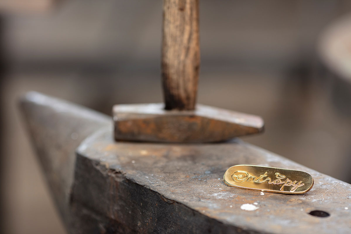 Close-up of a handcrafted brass tag engraved with 'OnEntropy', placed on a worn anvil beside a wooden-handled hammer—symbolizing artisanal identity and craftsmanship